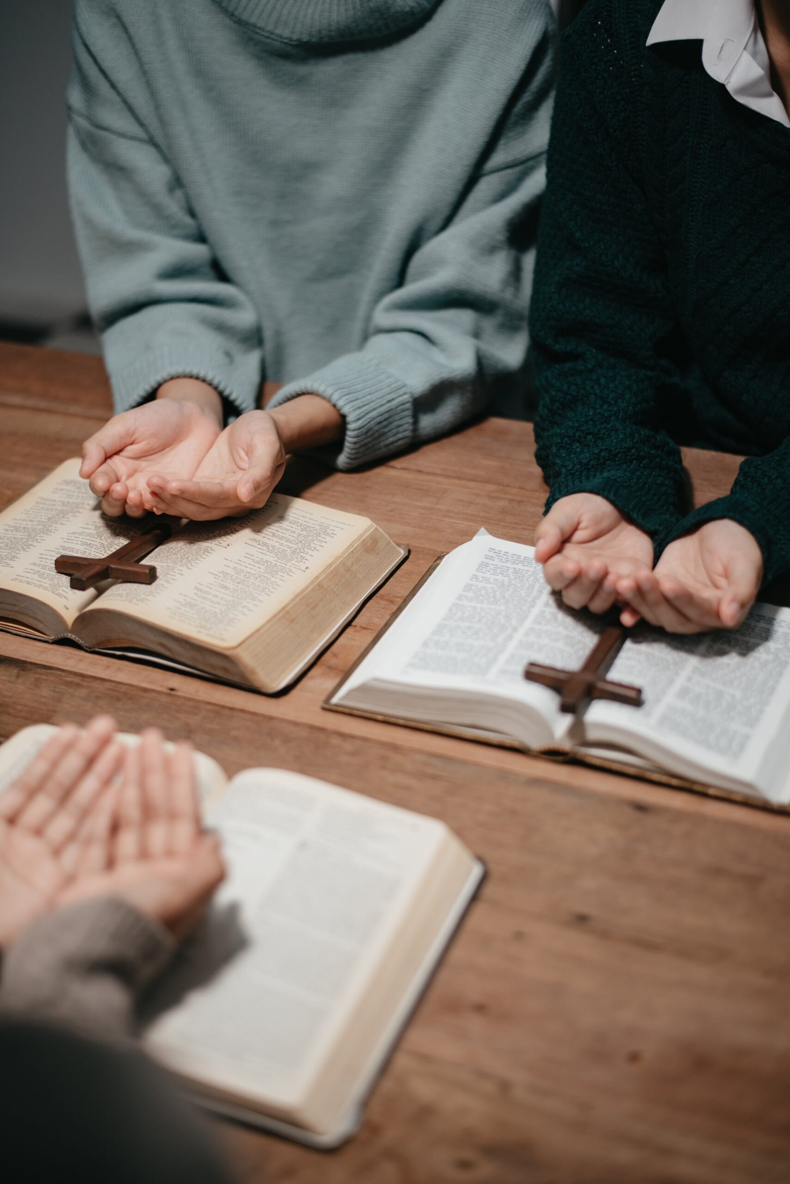 Group of Christians sit together and pray around a wooden table with blurred open Bible pages in their homeroom. Prayer for brothers, faith, hope, and seek the blessings of God.
